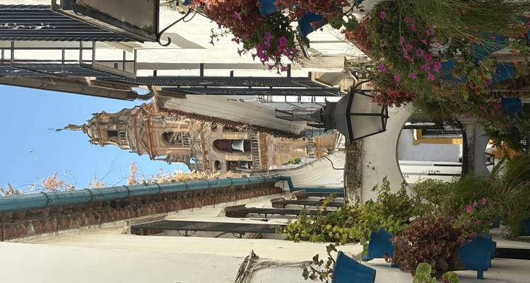 Une ruelle étroite avec des pots de fleurs colorés et un clocher d'église en arrière-plan.