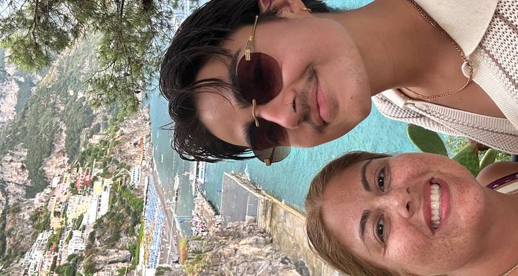 Two people posing with a scenic view of Positano coastline in the background.