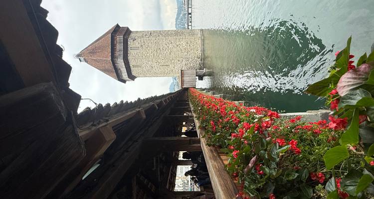 A stone tower with flowers along a covered wooden bridge over water.