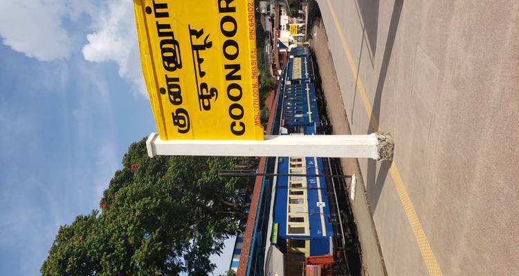 A train station with a sign for Coonoor and a blue train on the platform.