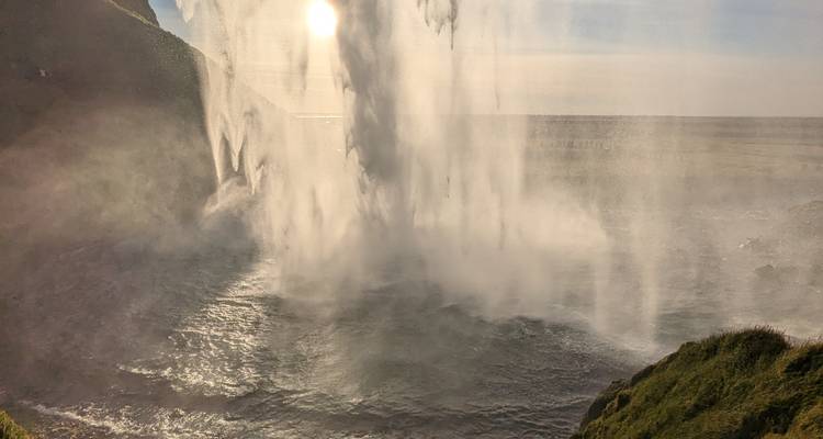 Uitzicht van achter een waterval met zonlicht dat door het water straalt.