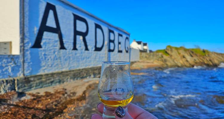 Verre de whisky tenu devant le panneau Ardbeg au bord de l'eau.
