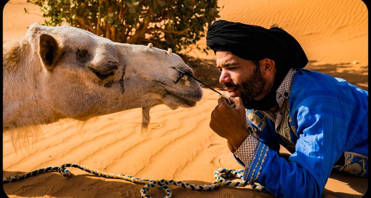 Man in traditional attire with a camel in the desert.