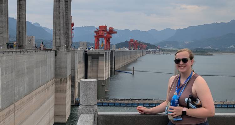 Una persona posando cerca de una presa con montañas de fondo.