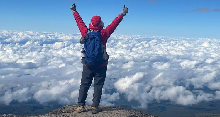 Randonneur face aux nuages, debout sur un sommet de montagne avec les bras levés.