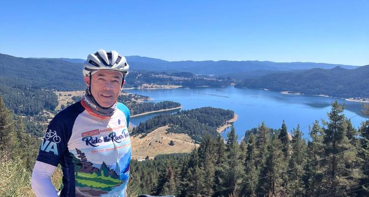 Man posing with bicycle against a scenic backdrop of a lake and mountains.