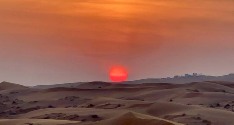 Atardecer sobre dunas del desierto con un edificio en el horizonte.