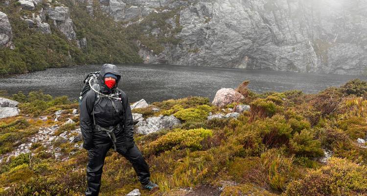 Excursionista de pie junto a un lago brumoso en un paisaje rocoso.