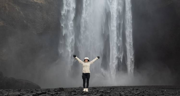 Persoon die staat met opgeheven armen voor een grote waterval.