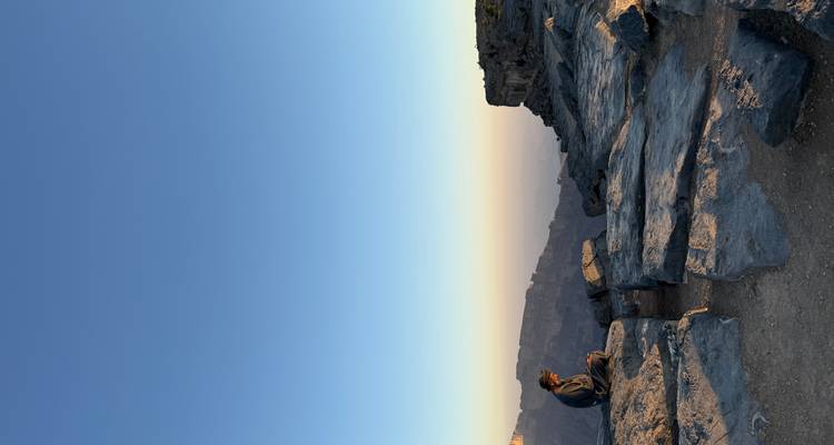 Persona sentada en una colina rocosa con amplia vista de montañas durante el atardecer.