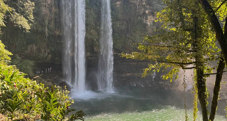 Dos cascadas altas con personas cerca de la base, rodeadas de bosque denso.