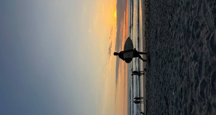 Surfer, der bei Sonnenuntergang entlang einem Strand geht, mit Menschen im Hintergrund.