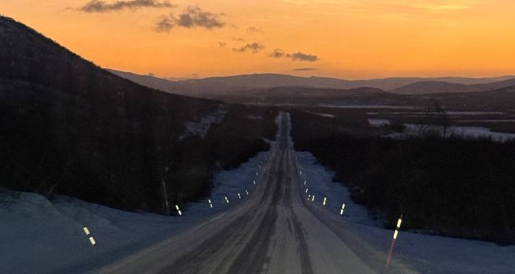 Carretera nevada que conduce hacia un atardecer con montañas en la distancia.