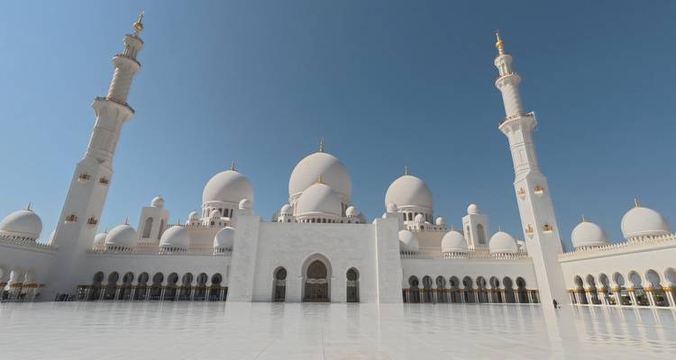 Pristine white domes and minarets of the Sheikh Zayed Grand Mosque reflected on its marble courtyard under a blue sky.