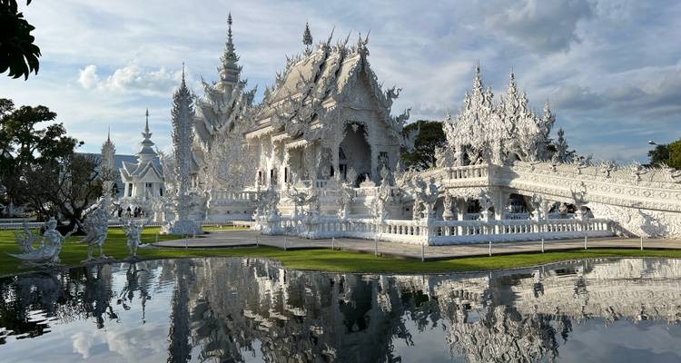 Dazzling white temple Wat Rong Khun gleams under bright sun, its reflection mirrored in a tranquil pond.