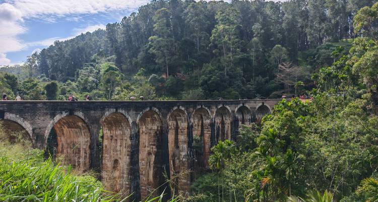 El histórico puente ferroviario de Nueve Arcos se extiende sobre un exuberante desfiladero de la selva con pequeños turistas caminando por su parte superior.