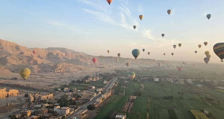 Dozens of colourful hot-air balloons float above green fields and ancient temples near Luxor at dawn.