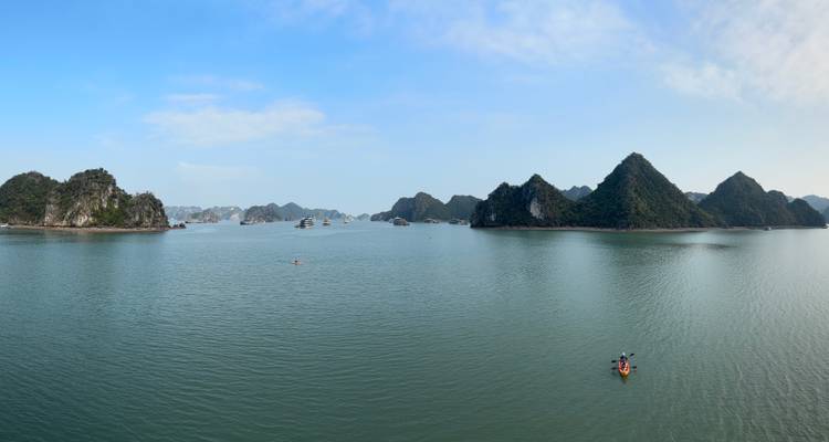 Vue panoramique d'eaux émeraude parsemées d'îles calcaires sous un ciel bleu éclatant, petit kayak au premier plan.