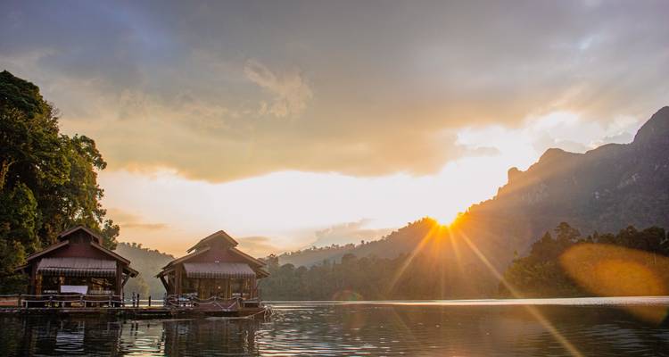 Des maisons flottantes sur radeau reposent sur un lac lisse comme un miroir dans le parc national de Khao Sok tandis que le soleil levant projette ses rayons sur les montagnes couvertes de jungle brumeuse.