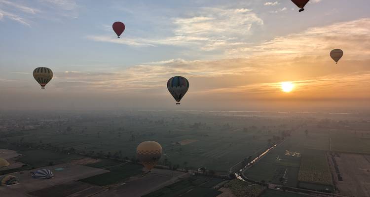 Globos aerostáticos coloridos flotan sobre campos verdes y el Valle del Nilo al amanecer, el sol dorado iluminando la bruma.