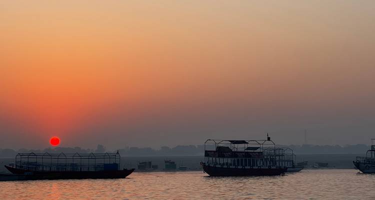 Barcos en el Ganges silueteados al amanecer con cielo naranja y agua tranquila.