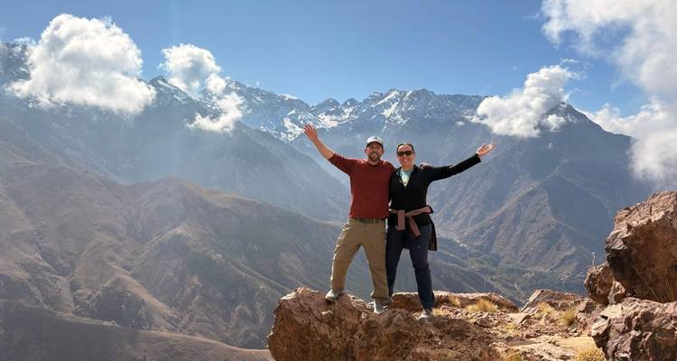 Excursionistas celebrando en la cima rocosa con los picos nevados del Alto Atlas detrás