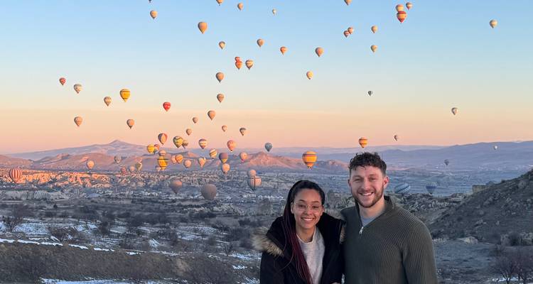 Couple souriant avec des dizaines de montgolfières colorées s'élevant au-dessus de la Cappadoce au lever du soleil.