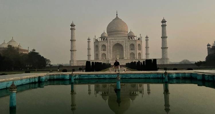 Das ikonische Taj Mahal spiegelt sich bei Morgendämmerung in einem stillen Wasserbecken wider, mit einem einsamen Besucher, der auf der zentralen Plattform sitzt.