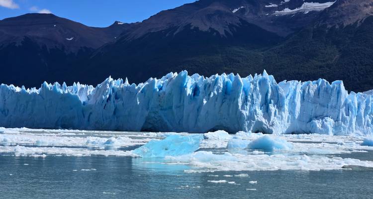 Dramatische blauwe ijsmuur van een gletsjer die uitkomt op een kalm meer met beboste bergen erachter
