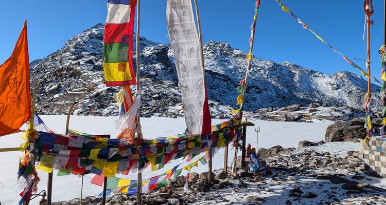 Drapeaux de prière colorés flottant au vent près d'un lac alpin gelé entouré de montagnes enneigées.