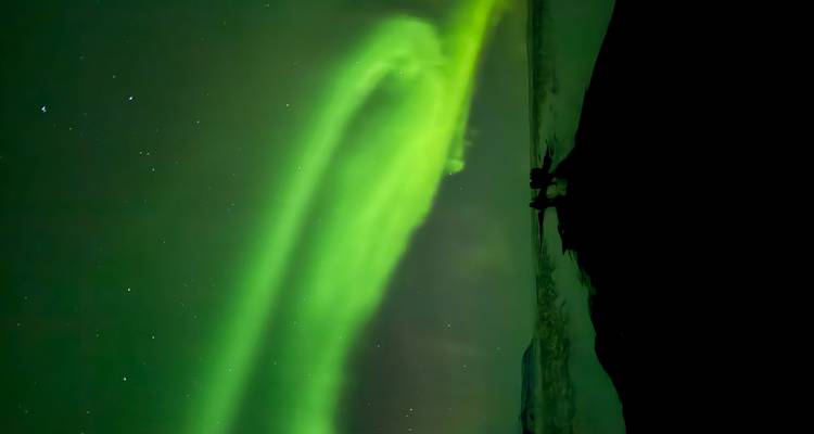 Vibrant green aurora dancing across the night sky above a dark snowy landscape with tiny silhouetted onlookers.