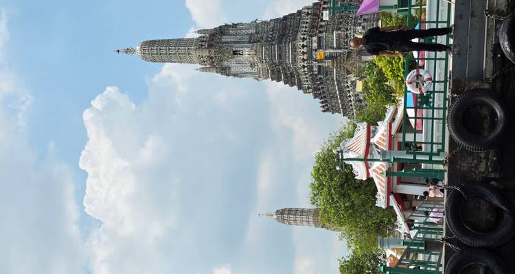 Le prang iconique du Wat Arun s'élève au-dessus d'un embarcadère au bord du fleuve sous un ciel bleu avec des nuages cotonneux et des visiteurs sur le quai
