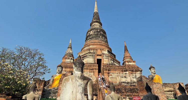 Vue grand-angle d'un majestueux chedi d'Ayutthaya flanqué de statues de Bouddha avec des visiteurs gravissant l'escalier central sous un ciel bleu dégagé