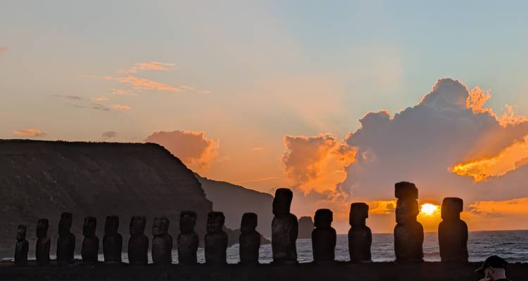 El sol sale entre las estatuas moai alineadas a lo largo de la costa, proyectando rayos a través de nubes dramáticas.