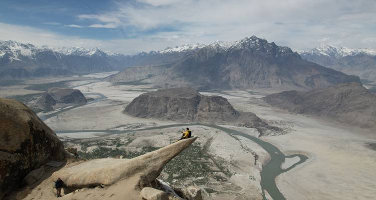 Des aventuriers assis sur un étroit affleurement rocheux très au-dessus d'une vallée de rivière sinueuse et de montagnes escarpées.