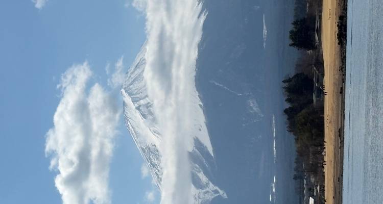 Le mont Fuji enneigé s'élève au-dessus de nuages bas avec un lac et un premier plan rural sous un ciel bleu.