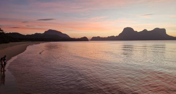 Espectacular atardecer rosa-anaranjado sobre una bahía tropical en calma con islas kársticas silueteadas en el horizonte y dos amigos de pie en la orilla del agua.