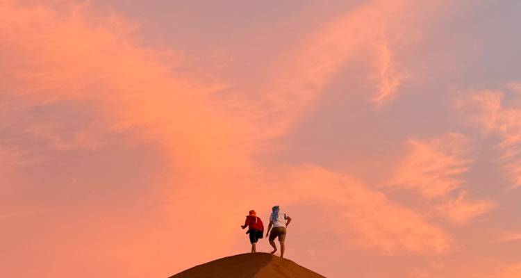 Twee kleine silhouetten beklimmen de kam van een zandduin tegen een dramatische roze-oranje zonsonderganghemel.