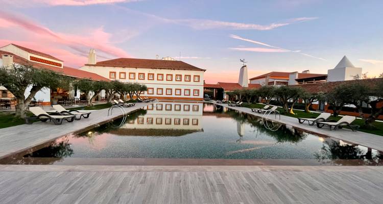 Complexe de luxe à la campagne au coucher du soleil avec piscine à débordement symétrique, chaises longues et oliviers reflétant les ciels pastel.