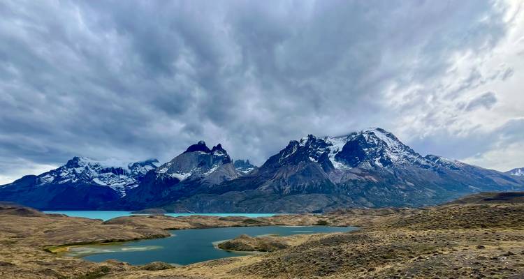 Dramáticas montañas cubiertas de nieve se alzan sobre lagos turquesa y estepas ondulantes bajo nubes melancólicas en la Patagonia.