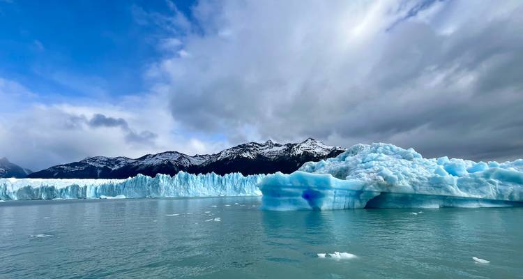 Una vasta pared de hielo azul dentado se eleva sobre un lago glacial en calma con montañas nevadas en la distancia.