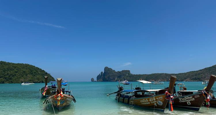 Des bateaux colorés à longue queue flottent sur une eau turquoise cristalline avec des îles calcaires au loin.