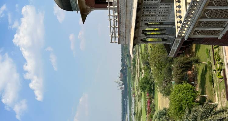 Vue panoramique depuis le balcon d'un fort historique donnant sur des plaines verdoyantes et le Taj Mahal au loin sous un ciel bleu parsemé de nuages