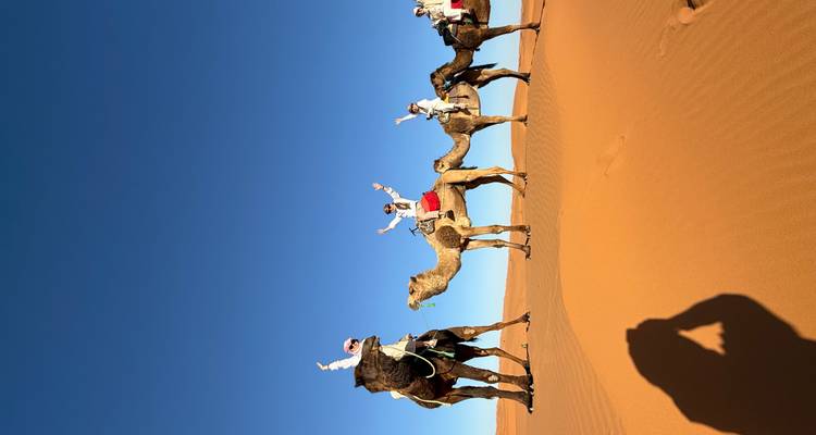 Caravana de camellos de viajeros levantando los brazos mientras cabalgan a través de imponentes dunas naranjas bajo un cielo azul profundo