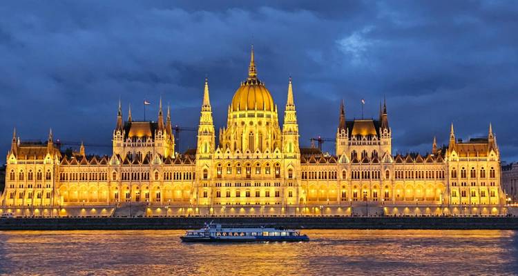 Bâtiment du Parlement hongrois brillamment illuminé la nuit de l'autre côté du Danube avec un bateau de croisière éclairé au premier plan.