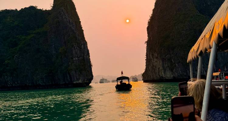 Bateau naviguant entre d'imposantes falaises calcaires au coucher de soleil orangé se reflétant sur une eau émeraude