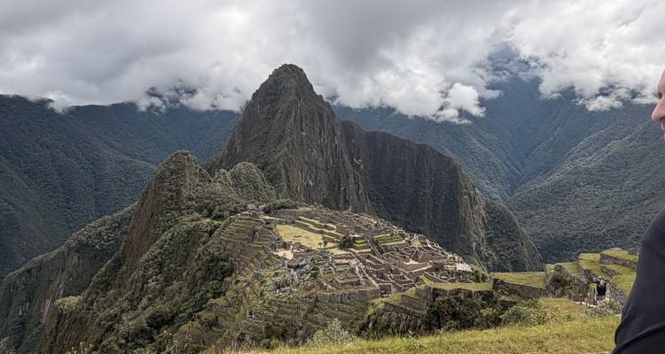 Vista icónica de la ciudadela inca de Machu Picchu rodeada de montañas verdes empinadas bajo un cielo nublado, con parte de un visitante visible en el borde.