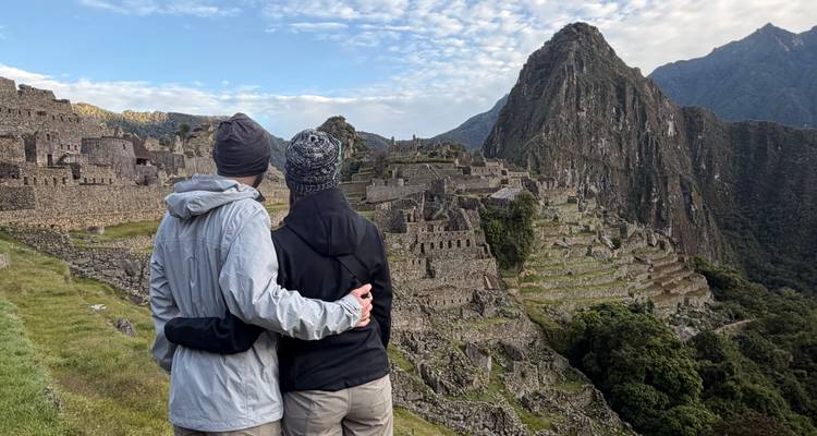 Una pareja vestida con equipo de senderismo se abraza mientras admira las terrazas de piedra de Machu Picchu al amanecer.