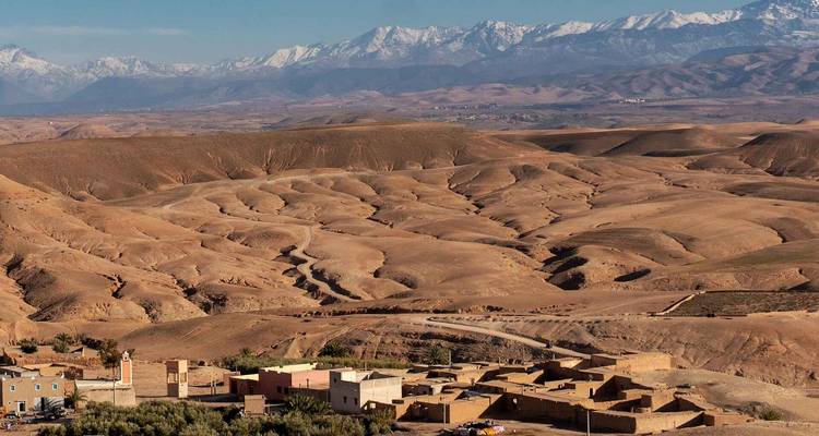 Panoramic view of arid rolling hills and a small Berber village set against the distant snow-capped Atlas Mountains.
