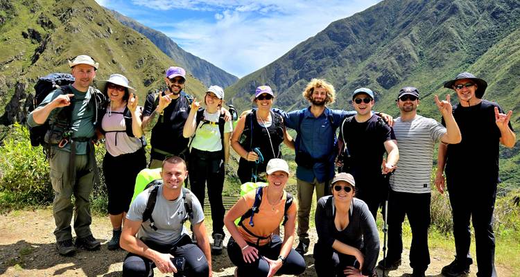 Glückliche Trekkinggruppe posiert gemeinsam auf dem Inka-Trail mit tiefgrünen Tälern und Gipfeln, die sich hinter ihnen unter blauem Himmel erstrecken.